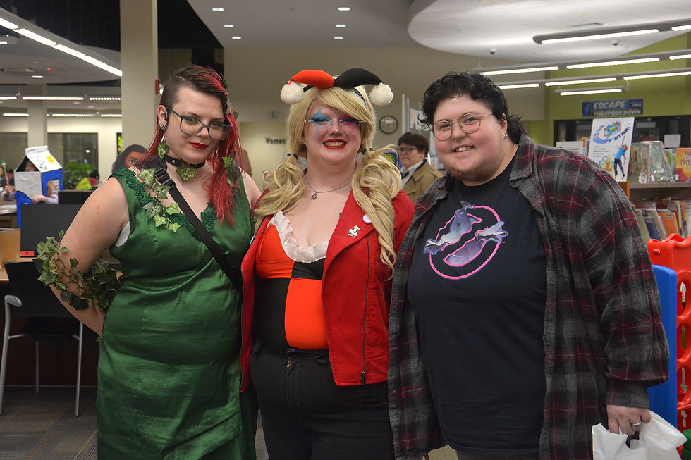 Three people in cosplay pose smiling in a library. One wears a green dress, another a red jacket, and the third a plaid shirt.