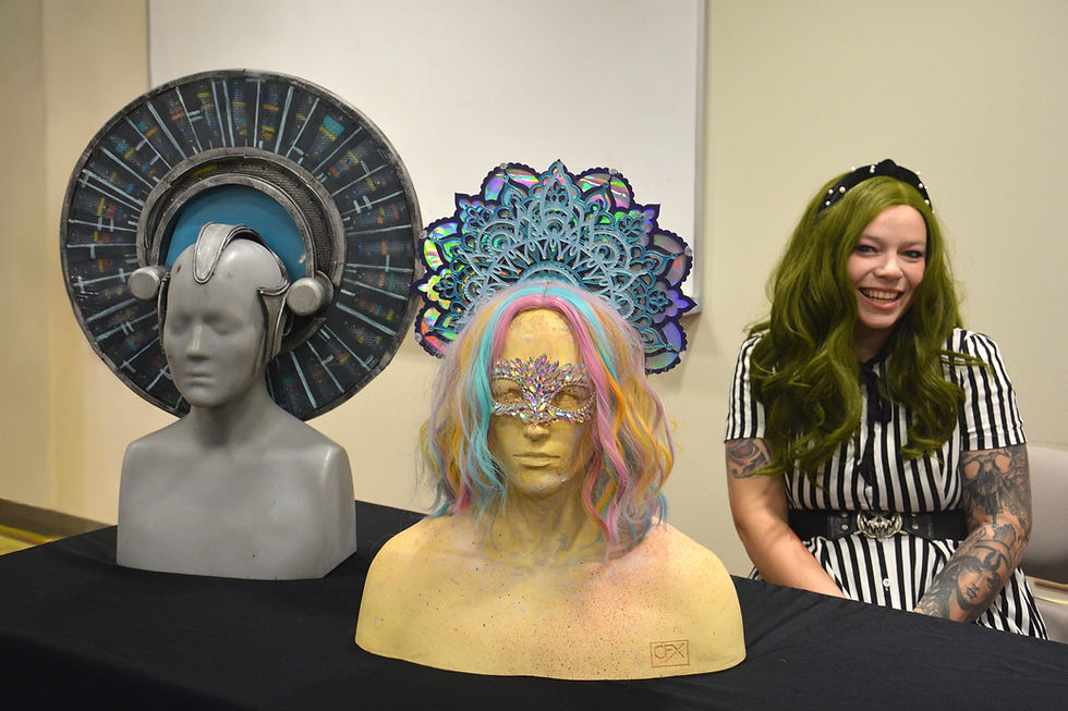 A person with green hair smiles next to two mannequin heads with intricate headdresses and masks. One has rainbow hair, set against a plain background.
