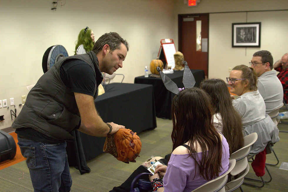 Man showing a pumpkin head to a girl with bunny ears. Seated audience in a room with decorated tables. Warm, interactive mood.