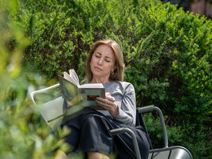 Woman reading a book in a chair amid lush green bushes, looking calm and relaxed outdoors.