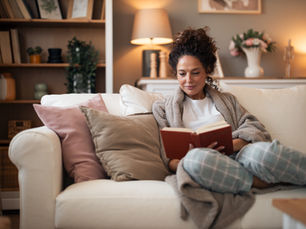 Woman in cozy robe reading a book on a white sofa, surrounded by pillows. Warm lighting, bookshelves, and flowers in soft, neutral tones.