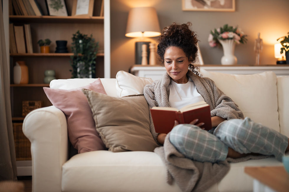 Woman in cozy robe reading a book on a white sofa, surrounded by pillows. Warm lighting, bookshelves, and flowers in soft, neutral tones.