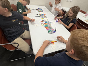 Four children color paper cutouts with markers around a classroom table, focused and quiet, with rows of colorful markers on the table.