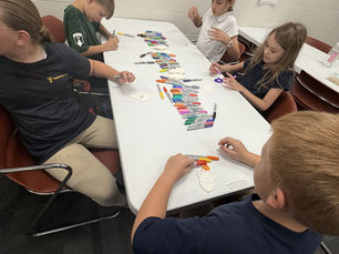 Four children color paper cutouts with markers around a classroom table, focused and quiet, with rows of colorful markers on the table.