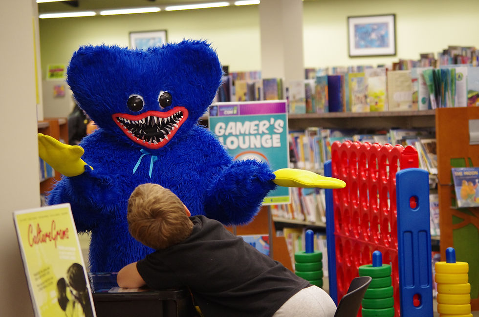 A child interacts with a person in a blue furry costume with sharp teeth in a library. Nearby, a "Gamer's Lounge" sign and colorful games.