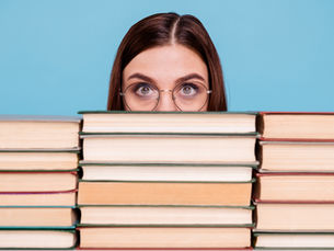 Woman with glasses peers over a stack of books against a blue background, conveying curiosity and focus.
