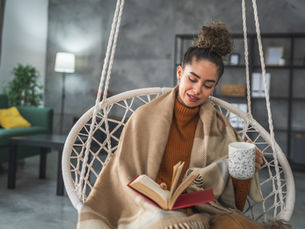 Woman reads a book while holding a mug in a cozy, netted swing. She's wrapped in a tan blanket, with a peaceful living room backdrop.