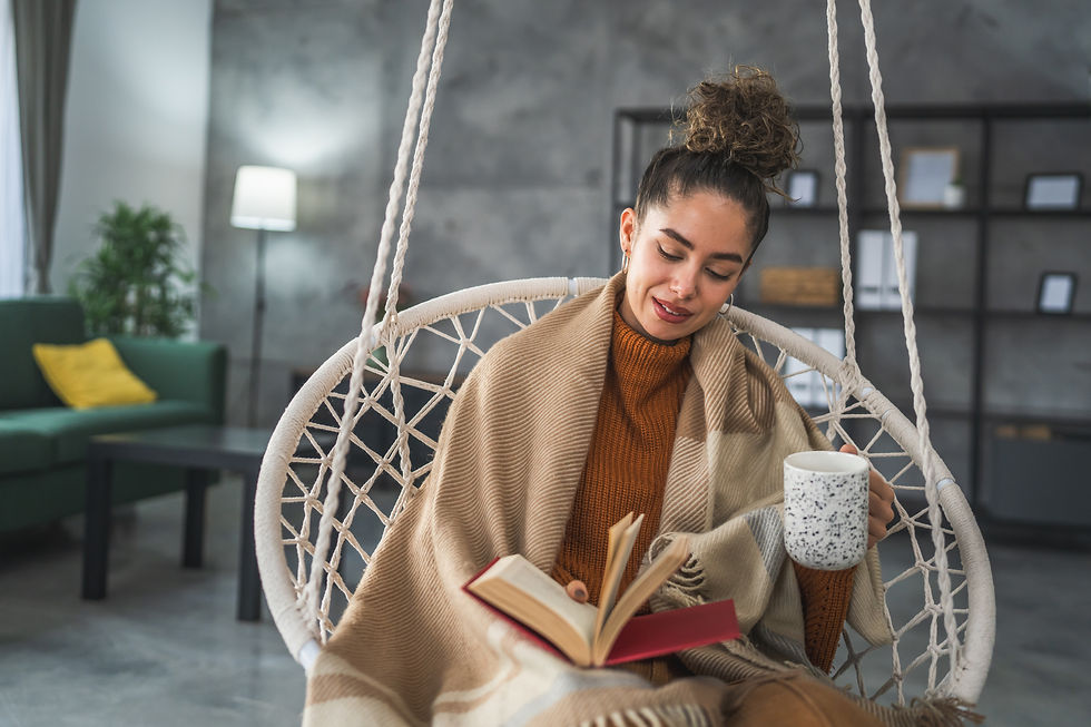 Woman reads a book while holding a mug in a cozy, netted swing. She's wrapped in a tan blanket, with a peaceful living room backdrop.