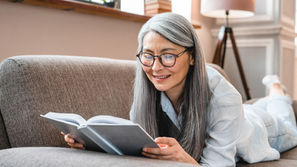Smiling older woman with gray hair and glasses reads a book on a sofa in a bright cozy living room.