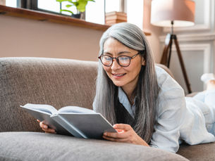 Smiling older woman with gray hair and glasses reads a book on a sofa in a bright cozy living room.