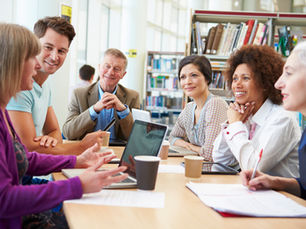 Diverse group smiles during a meeting around a table with laptops and papers in a bright library, bookshelves behind them.