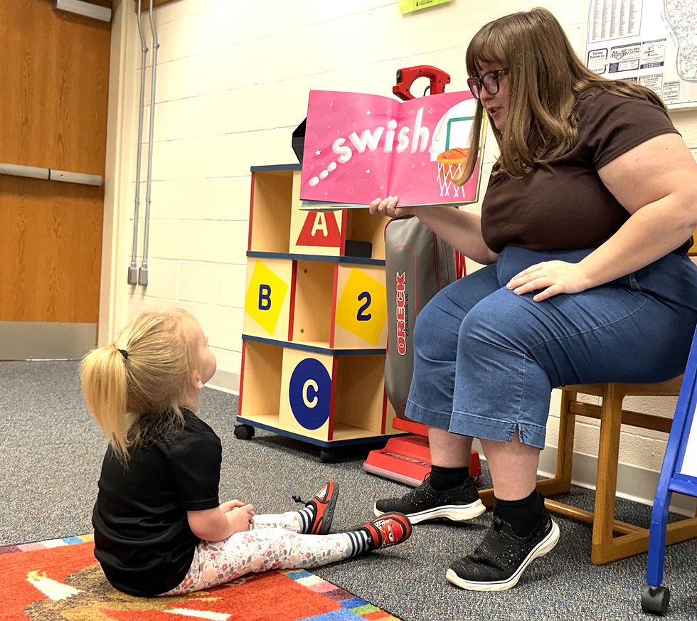 A woman shows a book reading "Swish" to a child in a classroom. The child sits on a colorful mat, looking up attentively.