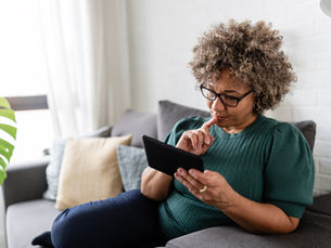 Woman with curly hair in green sweater sits on a couch, focusing on a tablet. Neutral mood, pillows and plant in background.