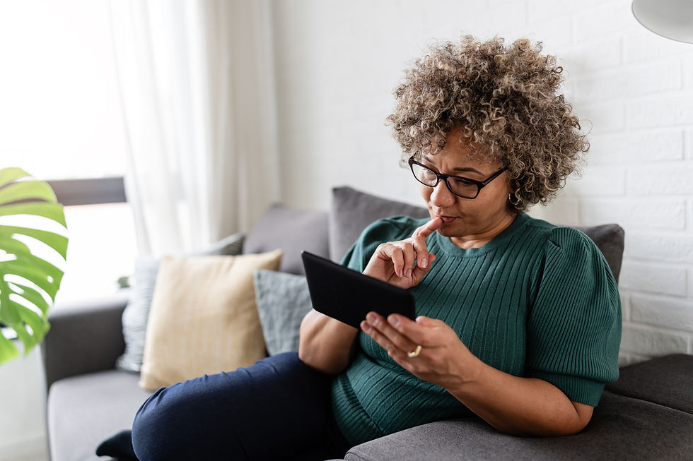 Woman with curly hair in green sweater sits on a couch, focusing on a tablet. Neutral mood, pillows and plant in background.