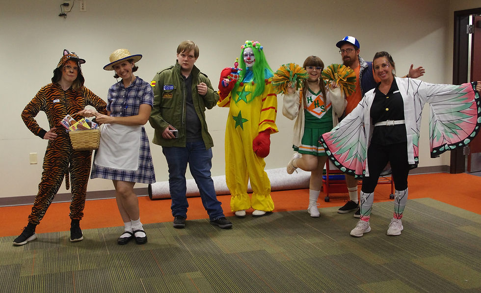 Seven people in costumes pose indoors on a green and orange carpet. Outfits include a tiger, clown, and cheerleader. Mood is festive.