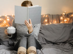 Person in cozy sweater reads a book on a bed with fairy lights, holding a mug of marshmallows. Relaxed, warm ambiance.
