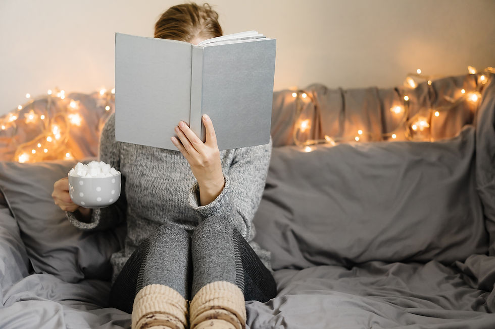 Person in cozy sweater reads a book on a bed with fairy lights, holding a mug of marshmallows. Relaxed, warm ambiance.