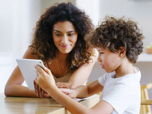 Mother and young boy in a bright kitchen look at a tablet together, smiling softly.