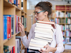 Woman in a polka dot blouse holding a stack of books, browses a library shelf. She wears glasses and appears focused. Bright, colorful books surround her.