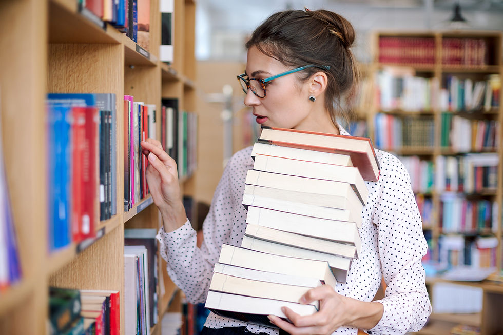 Woman in a polka dot blouse holding a stack of books, browses a library shelf. She wears glasses and appears focused. Bright, colorful books surround her.