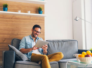 Man in glasses, smiling while using tablet on gray sofa in cozy room with wooden decor, bright light, and colorful fruit bowl. Relaxed mood.