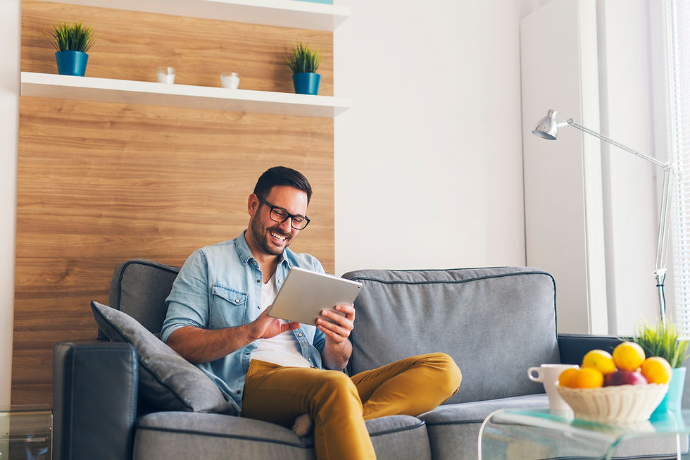 Man in glasses, smiling while using tablet on gray sofa in cozy room with wooden decor, bright light, and colorful fruit bowl. Relaxed mood.