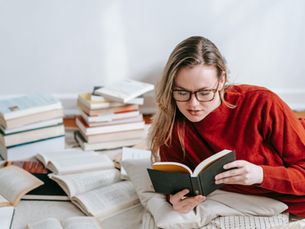 Woman in red sweater, glasses, reading a book on the floor with scattered books around her. Relaxed, cozy setting with white walls.