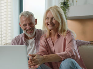 Smiling couple sitting on a couch, using a laptop. Bright room, plants in background. Both wearing pink shirts, conveying a joyful mood.