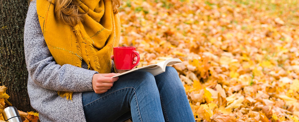 Person sitting on the autumn leaf covered ground under a tree holding a mug in one hand with a book on their lap