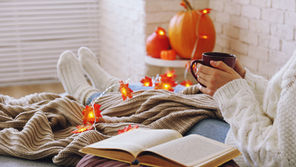 Cozy autumn scene of a person in a white sweater holding a mug, reading with an open book, pumpkins and glowing leaf lights nearby