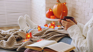 Cozy autumn scene of a person in a white sweater holding a mug, reading with an open book, pumpkins and glowing leaf lights nearby