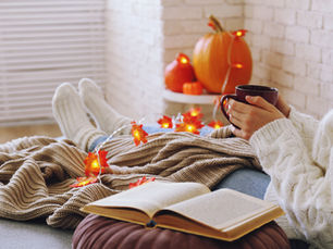 Cozy autumn scene of a person in a white sweater holding a mug, reading with an open book, pumpkins and glowing leaf lights nearby
