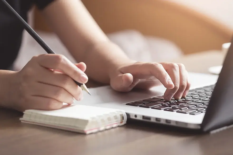 Hands writing in a notebook with a pencil and typing on a laptop. Soft lighting, wooden desk, focused and productive setting.